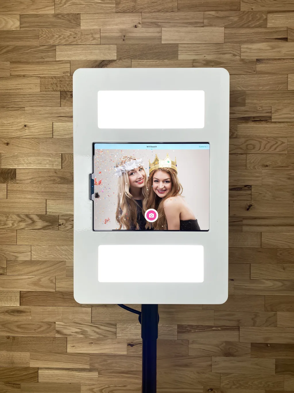 Design-R-Music Fotobox mit Stativ vor warmer Holzwand, Touchscreen zeigt zwei lachende Gäste mit Pappaccessoires
