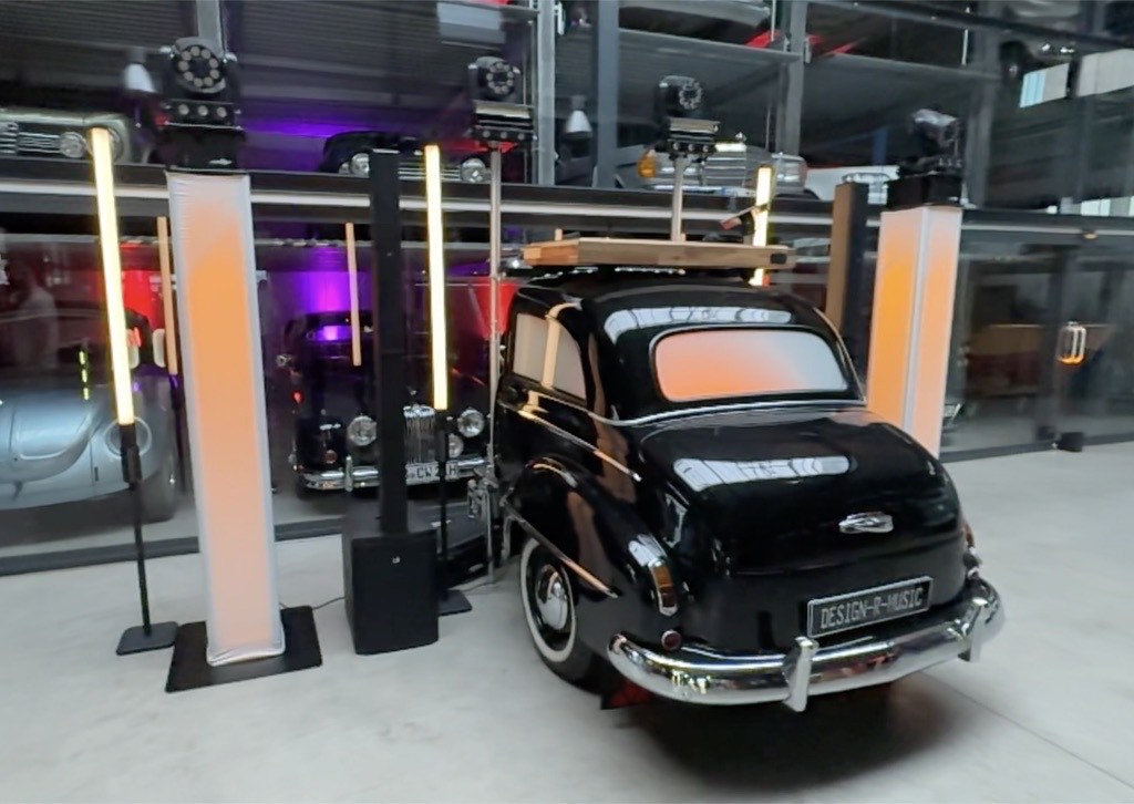 DJ-Setup mit warm beleuchteten Sound-Towern und Pioneer-Pult vor schwarzem Oldtimer mit DESIGN-R-MUSIC-Kennzeichen in der Oldtimer Garage Paderborn DJ-Setup mit warm beleuchteten Sound-Towern und Pioneer-Pult vor schwarzem Oldtimer mit DESIGN-R-MUSIC-Kennzeichen in der Oldtimer Garage Paderborn