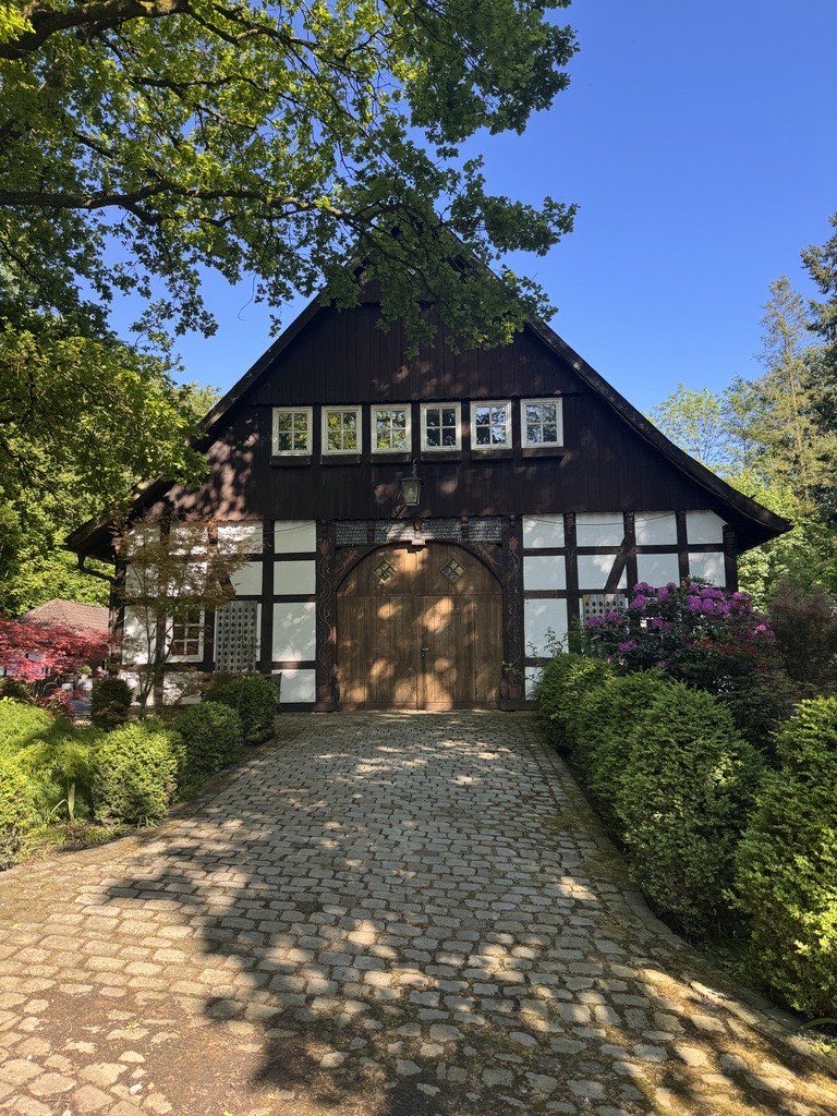 Historisches Fachwerkhaus Waterbör in Bielefeld-Senne mit Holztür, Kopfsteinpflaster-Auffahrt und blühenden Rhododendren unter blauem Himmel Historisches Fachwerkhaus Waterbör in Bielefeld-Senne mit Holztür, Kopfsteinpflaster-Auffahrt und blühenden Rhododendren unter blauem Himmel